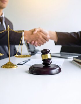 Two business people shaking hands over a legal contract with a wooden gavel and golden scales of justice on a white desk in an office setting.