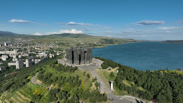 Drone shot capturing the massive bronze and stone pillars of the Chronicle of Georgia monument complex, situated on a green hill overlooking the vast Tbilisi Sea reservoir under a clear blue sky