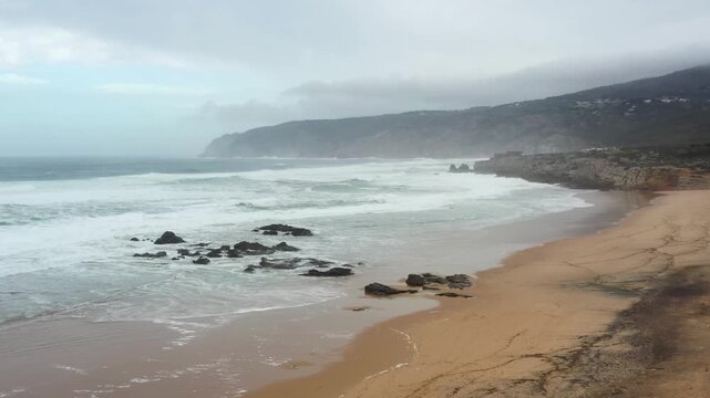 Waves crash against the rocks at Guincho Beach as clouds linger overhead. The golden sand glimmers, revealing nature's power and tranquility after a storm.