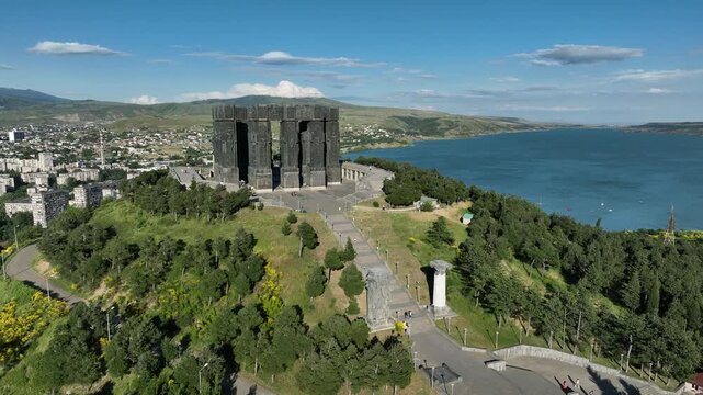 Drone shot capturing the massive bronze and stone pillars of the Chronicle of Georgia monument complex, situated on a green hill overlooking the vast Tbilisi Sea reservoir under a clear blue sky
