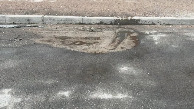 Worn asphalt road with potholes showing curbside emergency lane, gravel shoulder, standing puddles, cracked surface, localized