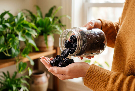 Woman gently pouring dark dried plums from a clear glass jar into her hand inside a bright room