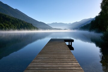 Obraz premium Serene wooden pier extending into a calm misty mountain lake surrounded by lush forests under a clear early morning sky