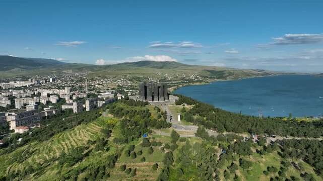 Drone shot capturing the massive bronze and stone pillars of the Chronicle of Georgia monument complex, situated on a green hill overlooking the vast Tbilisi Sea reservoir under a clear blue sky