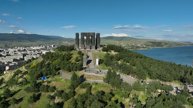 Drone shot capturing the massive bronze and stone pillars of the Chronicle of Georgia monument complex, situated on a green hill overlooking the vast Tbilisi Sea reservoir under a clear blue sky