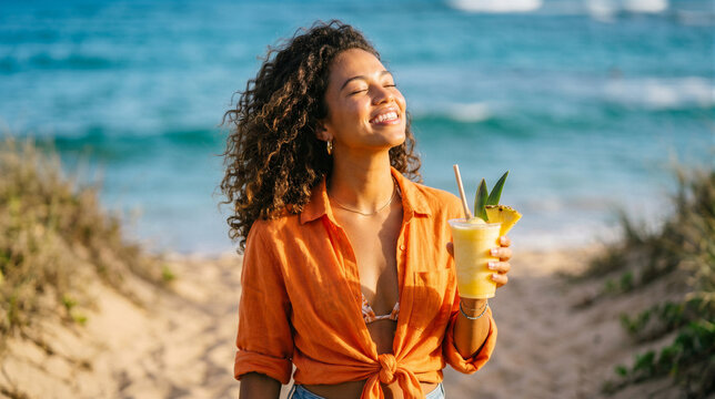 Happy woman with curly hair holding a pineapple smoothie on a sunny beach, enjoying a tropical vacation in a bright orange shirt