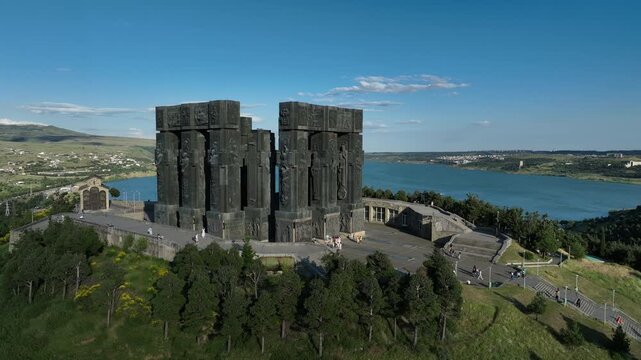 Drone shot capturing the massive bronze and stone pillars of the Chronicle of Georgia monument complex, situated on a green hill overlooking the vast Tbilisi Sea reservoir under a clear blue sky