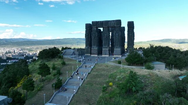 Drone shot capturing the massive bronze and stone pillars of the Chronicle of Georgia monument complex, situated on a green hill overlooking the vast Tbilisi Sea reservoir under a clear blue sky