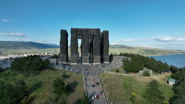 Drone shot capturing the massive bronze and stone pillars of the Chronicle of Georgia monument complex, situated on a green hill overlooking the vast Tbilisi Sea reservoir under a clear blue sky