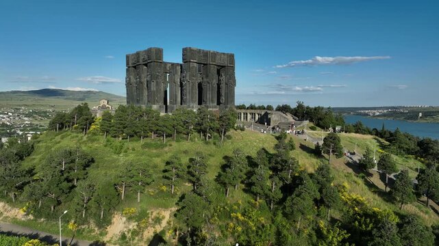 Drone shot capturing the massive bronze and stone pillars of the Chronicle of Georgia monument complex, situated on a green hill overlooking the vast Tbilisi Sea reservoir under a clear blue sky