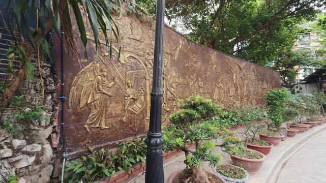 Ornate golden bas relief mural featuring an angel addressing mary in the annunciation, detailing various religious figures and biblical narratives along a long wall in a public garden and green park