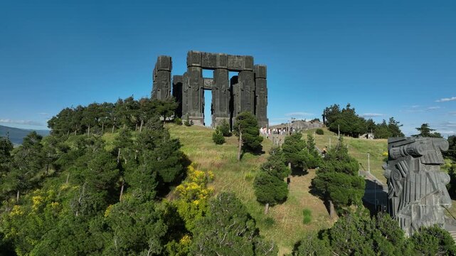 Drone shot capturing the massive bronze and stone pillars of the Chronicle of Georgia monument complex, situated on a green hill overlooking the vast Tbilisi Sea reservoir under a clear blue sky