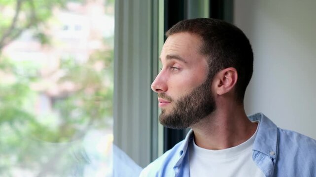 Pensive handsome young man with a beard thoughtfully looking out the window, contemplating and reflecting while observing the view from his home with a serious and pensive expression