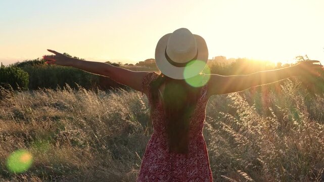 Back view of a young woman with long hair and a hat walking through a golden field, raising her arms to the warm sunset light and feeling a sense of freedom, joy, and connection with nature