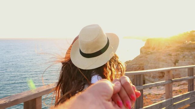 Beautiful woman with a sun hat leading her partner by the hand along a wooden boardwalk, smiling as they walk towards a breathtaking ocean and cliff view during a romantic golden sunset