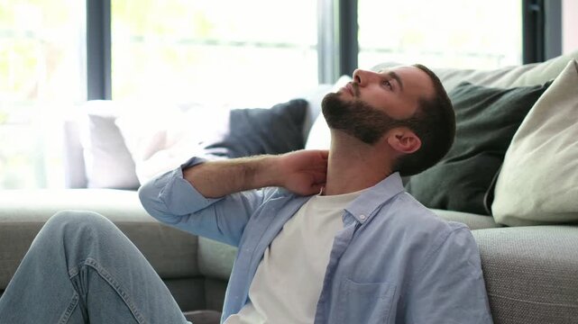 Stressed young man feeling tired and suffering from neck pain, massaging his tense muscles with his hand while sitting on the floor at home due to sedentary lifestyle or anxiety