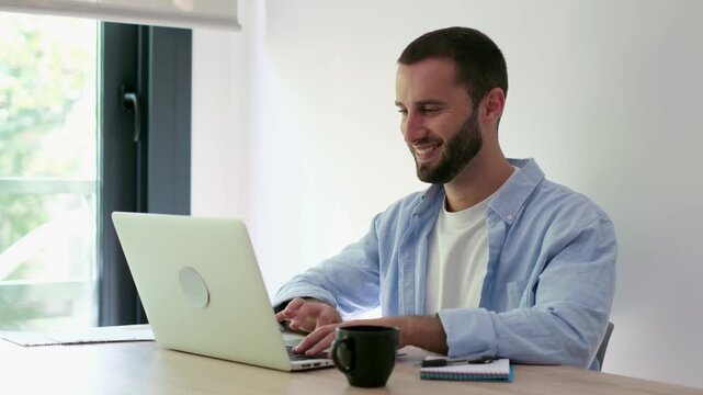 Millennial professional working from home, smiling and waving during an online conference call, multitasking on his notebook while sitting at his desk next to a bright window