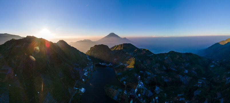 Aerial view of Telaga Cebong lake surrounded by terraced agricultural fields and mountain peaks at sunrise in Kejajar, Central Java, Indonesia.