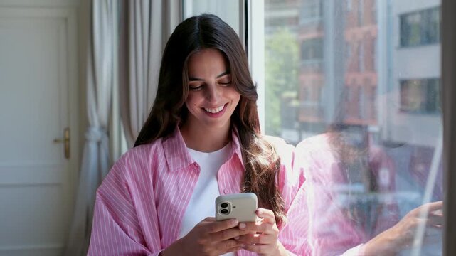 Beautiful young woman with long dark hair smiling while using her smartphone for texting and social media, standing by a large window with natural light in a cozy apartment setting
