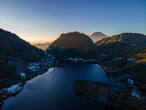 Aerial view of Telaga Cebong lake surrounded by terraced agricultural fields and hills at sunrise in Kejajar, Central Java, Indonesia.