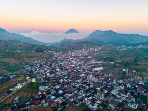 Aerial view of the Dieng plateau town of Kejajar nestled among terraced agricultural fields and volcanic mountains under a soft sunrise sky in Kejajar, Central Java, Indonesia.