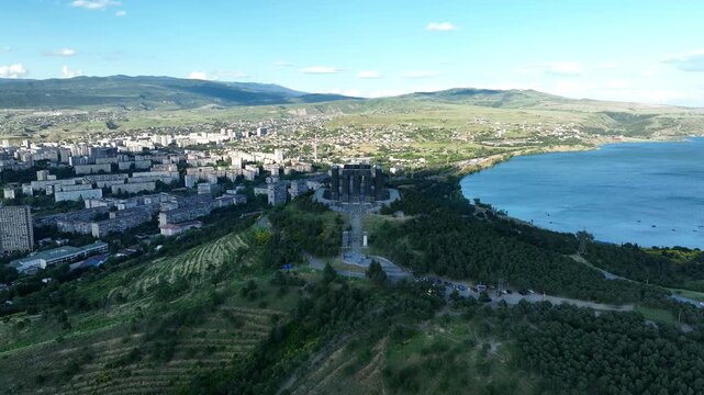Drone shot capturing the massive bronze and stone pillars of the Chronicle of Georgia monument complex, situated on a green hill overlooking the vast Tbilisi Sea reservoir under a clear blue sky