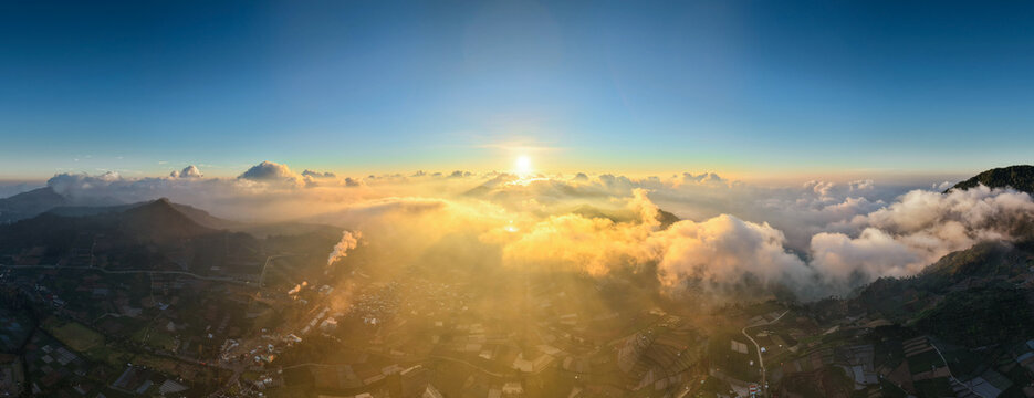 Aerial view of the sunrise over the Dieng Plateau with golden light rays illuminating clouds, terraced agricultural fields, and mountainous terrain in Kejajar, Central Java, Indonesia.