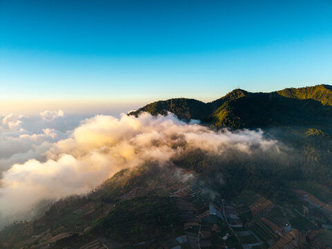 Aerial view of the Dieng plateau landscape with terraced agricultural fields partially covered by morning clouds under a clear blue sky in Kejajar, Central Java, Indonesia.