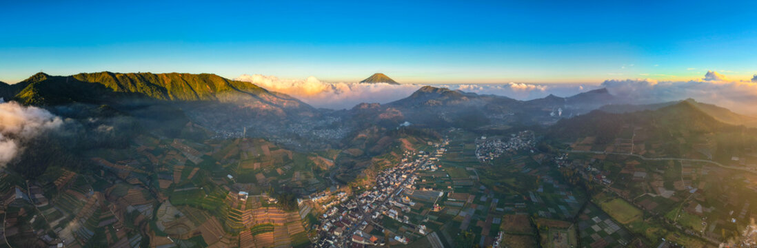 Aerial view of the Dieng plateau featuring lush terraced agricultural fields, volcanic mountains, and a town in the valley under a clear sky at sunrise in Kejajar, Central Java, Indonesia.