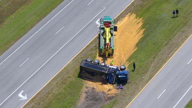 Tow truck lifting overturned trailer filled with spilled sand materials.