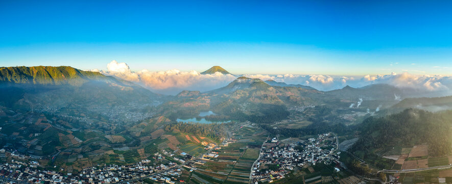 Aerial view of the Dieng Plateau landscape featuring lush green mountains, a serene lake, terraced agricultural fields, and a village nestled in the valley in Kejajar, Central Java, Indonesia.