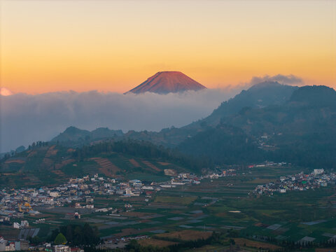 Aerial view of Dieng plateau featuring a volcano rising above clouds with terraced agricultural fields and a small town in Kejajar, Central Java, Indonesia.