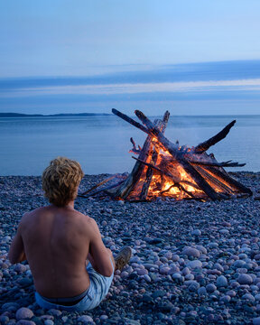 Person watches bonfire on Lake Superior beach vertical | A person sits before a glowing bonfire on a rocky beach along Lake Superior's north shore at dusk, in portrait orientation.
