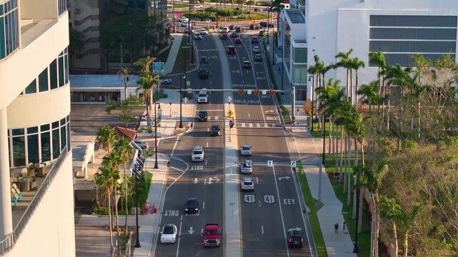 Crowded city street in Sarasota with people walking across marked crosswalk while cars wait at traffic light. Urban transportation and pedestrian safety concept.