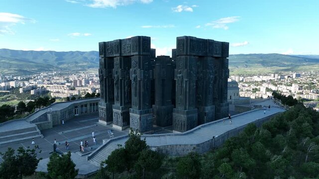 Drone shot capturing the massive bronze and stone pillars of the Chronicle of Georgia monument complex, situated on a green hill overlooking the vast Tbilisi Sea reservoir under a clear blue sky