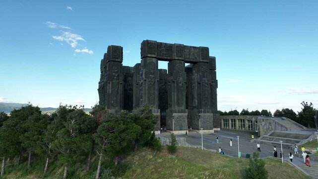 Drone shot capturing the massive bronze and stone pillars of the Chronicle of Georgia monument complex, situated on a green hill overlooking the vast Tbilisi Sea reservoir under a clear blue sky
