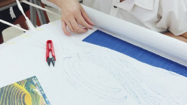Artisan's hands meticulously working on a large embroidery piece. Carefully stitching blue silk thread onto white fabric. Demonstrating precision and dedication to the craft of traditional needlework