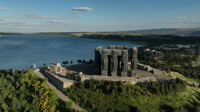Drone shot capturing the massive bronze and stone pillars of the Chronicle of Georgia monument complex, situated on a green hill overlooking the vast Tbilisi Sea reservoir under a clear blue sky