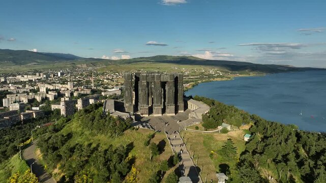 Drone shot capturing the massive bronze and stone pillars of the Chronicle of Georgia monument complex, situated on a green hill overlooking the vast Tbilisi Sea reservoir under a clear blue sky