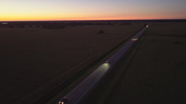 Overhead shot of two lane road stretching through open fields with cars driving forward with headlights on. Freedom and road trip adventure.