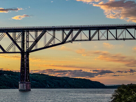 Golden hour light bathes the Hudson River as the Walkway Over the Hudson stretches across calm waters, framed by rolling hills and scattered clouds, capturing a serene Poughkeepsie evening view