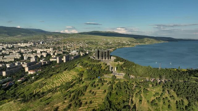 Drone shot capturing the massive bronze and stone pillars of the Chronicle of Georgia monument complex, situated on a green hill overlooking the vast Tbilisi Sea reservoir under a clear blue sky
