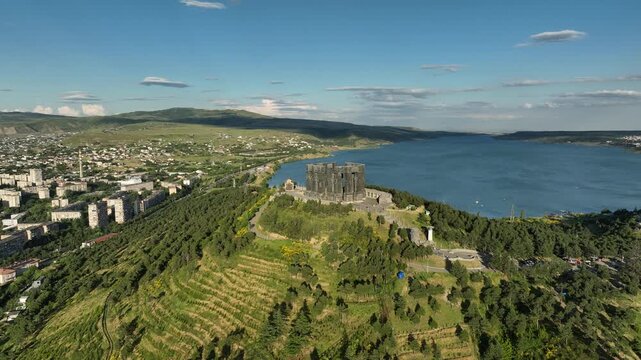 Drone shot capturing the massive bronze and stone pillars of the Chronicle of Georgia monument complex, situated on a green hill overlooking the vast Tbilisi Sea reservoir under a clear blue sky