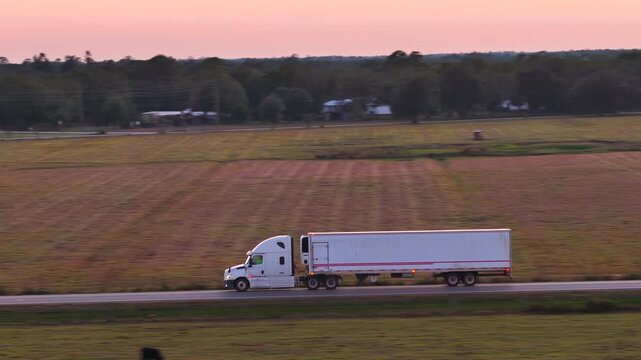 Two lane road through open fields with tractor trailer moving forward at golden hour. Cargo hauling and long distance delivery concept.