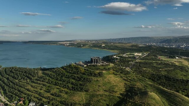 Drone shot capturing the massive bronze and stone pillars of the Chronicle of Georgia monument complex, situated on a green hill overlooking the vast Tbilisi Sea reservoir under a clear blue sky