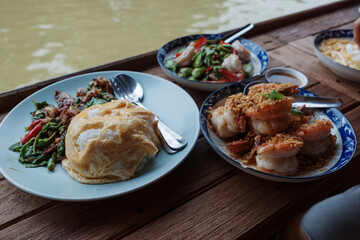 Fototapeta premium An eye-level photograph of a traditional Thai lunch set served on a wooden table beside a river, featuring a plate of rice topped with a creamy omelet alongside stir-fried shrimp dishes. 