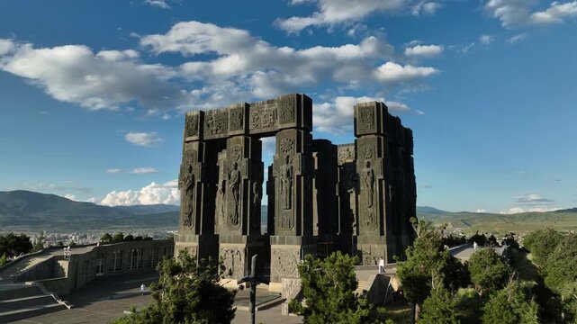 Drone shot capturing the massive bronze and stone pillars of the Chronicle of Georgia monument complex, situated on a green hill overlooking the vast Tbilisi Sea reservoir under a clear blue sky