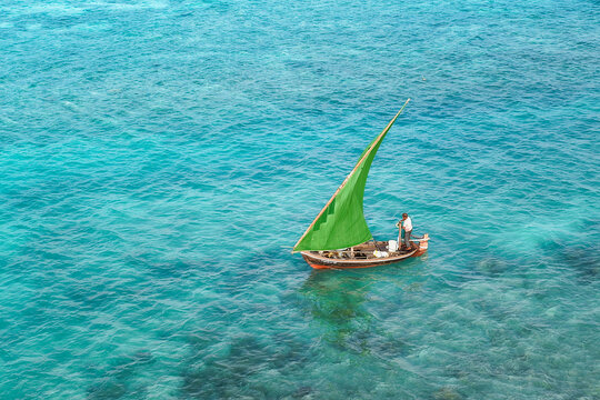 Aerial view of a traditional wooden boat with a vibrant green sail navigating the clear turquoise waters with a fisherman on board in Vaikaradhoo, Haa Dhaalu City, Maldives.