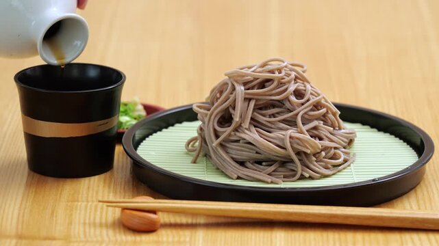 Pouring dashi tsuyu sauce into a cup for chilled zaru soba noodles on a dinner table, with sake.