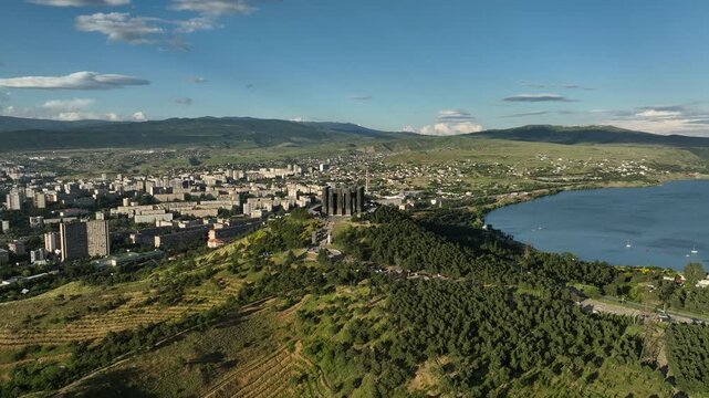 Drone shot capturing the massive bronze and stone pillars of the Chronicle of Georgia monument complex, situated on a green hill overlooking the vast Tbilisi Sea reservoir under a clear blue sky